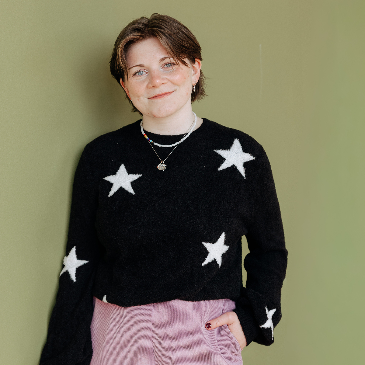 Person in a sweater with stars on it grinning at the camera against a green wall.