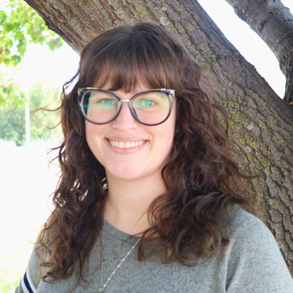 Person wearing glasses and smiling next to a tree outside.